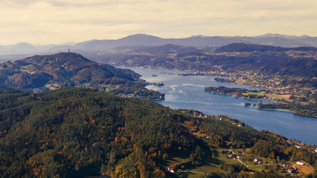 Herbstliche Luftaufnahme des Wörthersees mit sich langsam orange färbenden Wäldern. In der Ferne kann der Aussichtsturm am Pyramidenkogel erkannt werden.