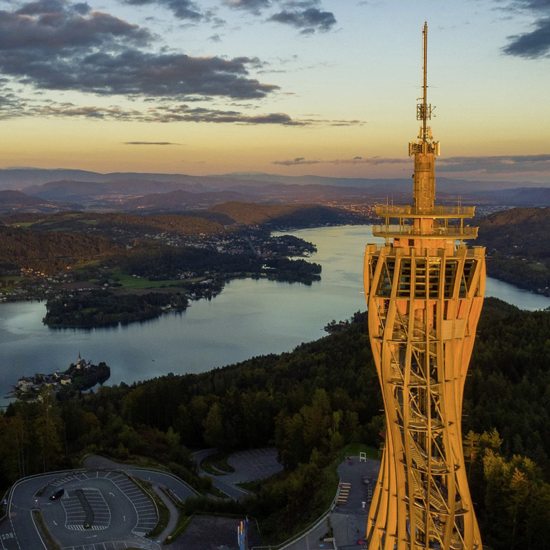 Nahaufnahme des Pyramidenkogel-Aussichtsturms in orangefarbenem Licht mit See und bewaldeten Hügeln im Hintergrund bei Sonnenuntergang