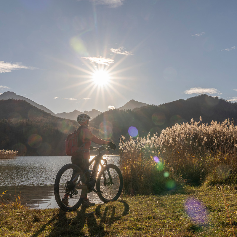 Person mit Fahrrad und Helm steht am Ufer eines Sees, im Hintergrund Berge und Sonne am klaren Himmel