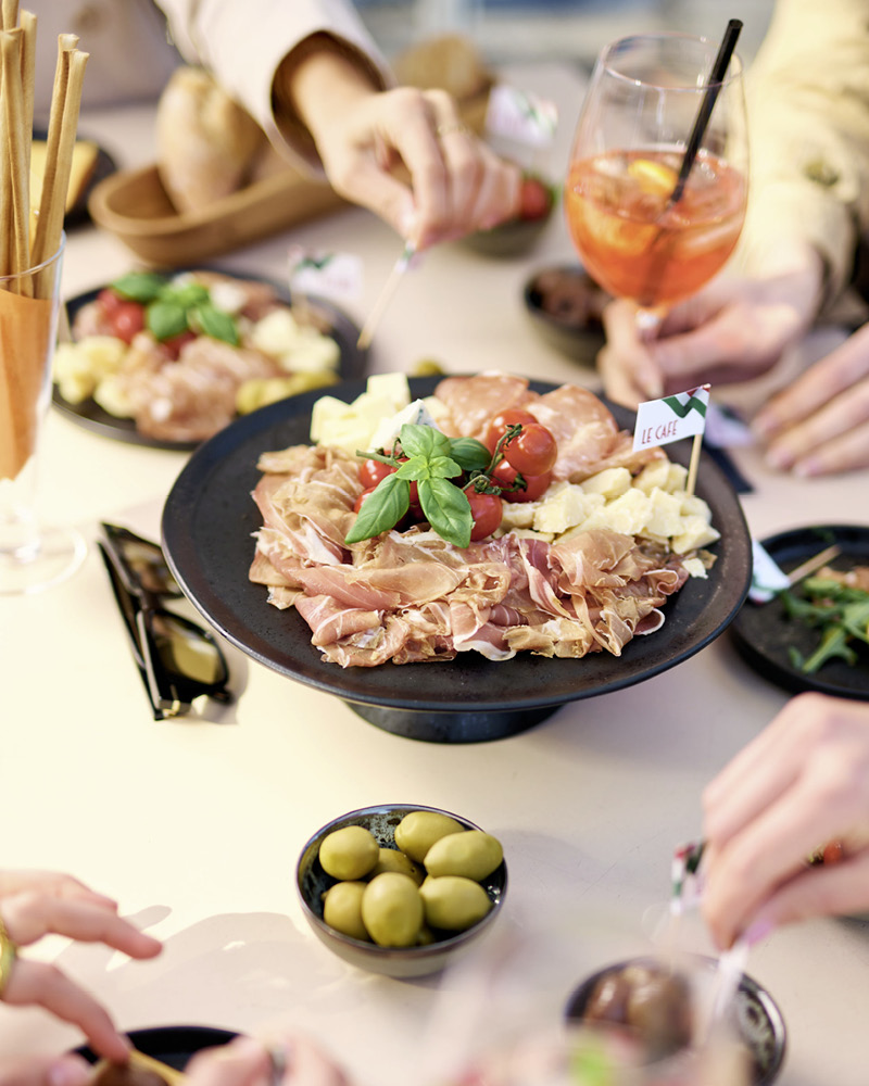 Vorspeisenteller mit Schinken, Käse, Tomaten und Basilikum, daneben Schale mit grünen Oliven, Hände greifen nach Essen, Glas mit orangem Getränk und Strohhalm