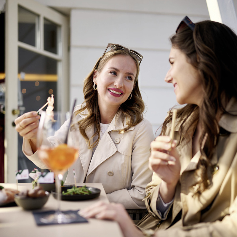 Zwei Frauen in beigen Trenchcoats sitzen an einem Tisch im Freien, eine hält eine Gabel mit Essen, auf dem Tisch stehen ein Glas mit orangefarbener Flüssigkeit und Teller mit Speisen.