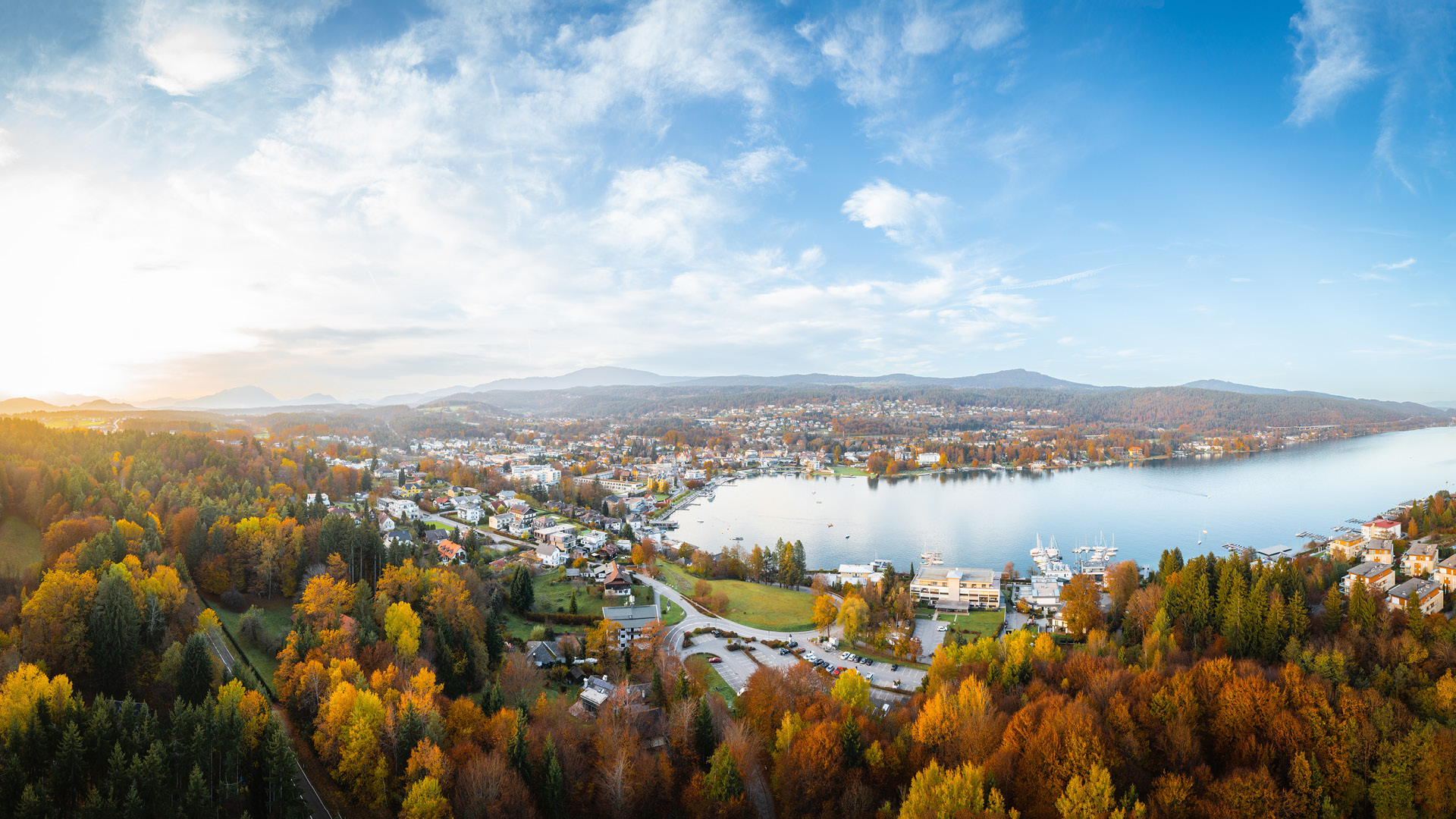 Luftaufnahme eines Sees mit umliegenden herbstlich gefärbten Wäldern und Wohngebieten unter bewölktem Himmel.